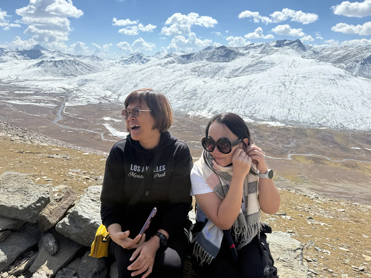 Two foreign female tourists sitting on a rock laughing with a distant river and snow-capped mountains near Babusar Top