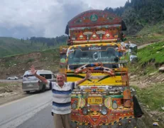 Foreign male tourist posing joyfully with raised arms in front of a colorful decorated Pakistani truck on a mountain road in Naran