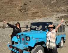 Two foreign female tourists posing with a local 4x4 Jeep amidst rugged brown mountains in Naran Kaghan